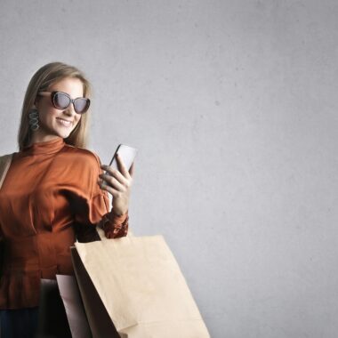 Smiling female in trendy wear and stylish sunglasses standing with paper shopping bags on background of gray wall and browsing mobile phone